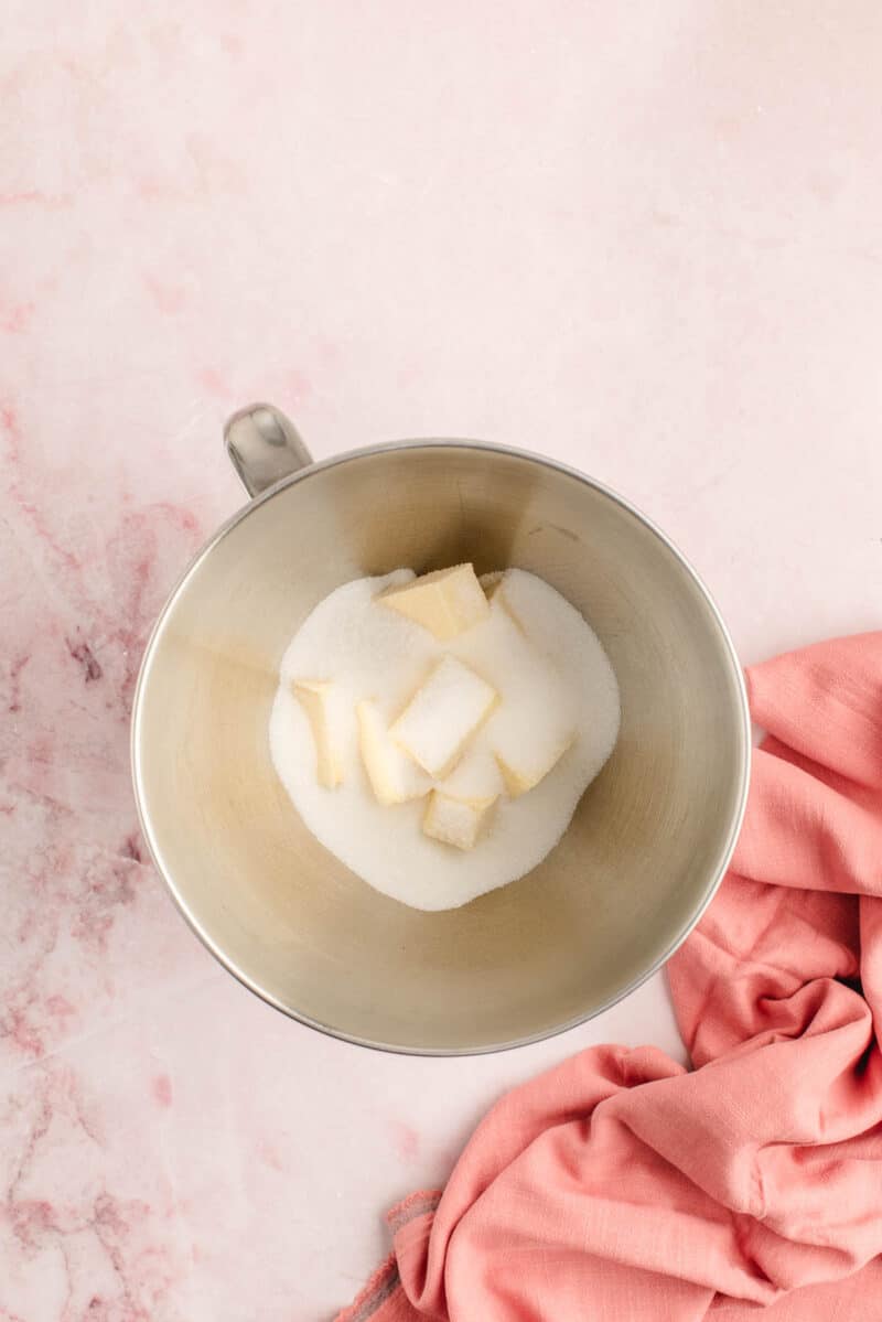 A metal mixing bowl with sugar and cubed butter inside, placed on a pink surface next to a pink cloth.