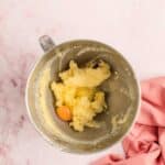 A metal mixing bowl with creamed butter and sugar, plus a cracked raw egg, sits on a pink surface next to a pink cloth.