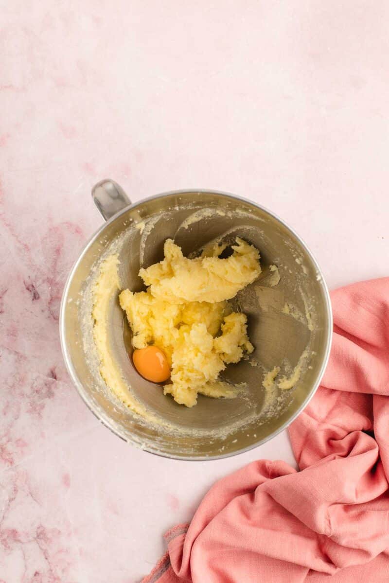 A metal mixing bowl with creamed butter and sugar, plus a cracked raw egg, sits on a pink surface next to a pink cloth.
