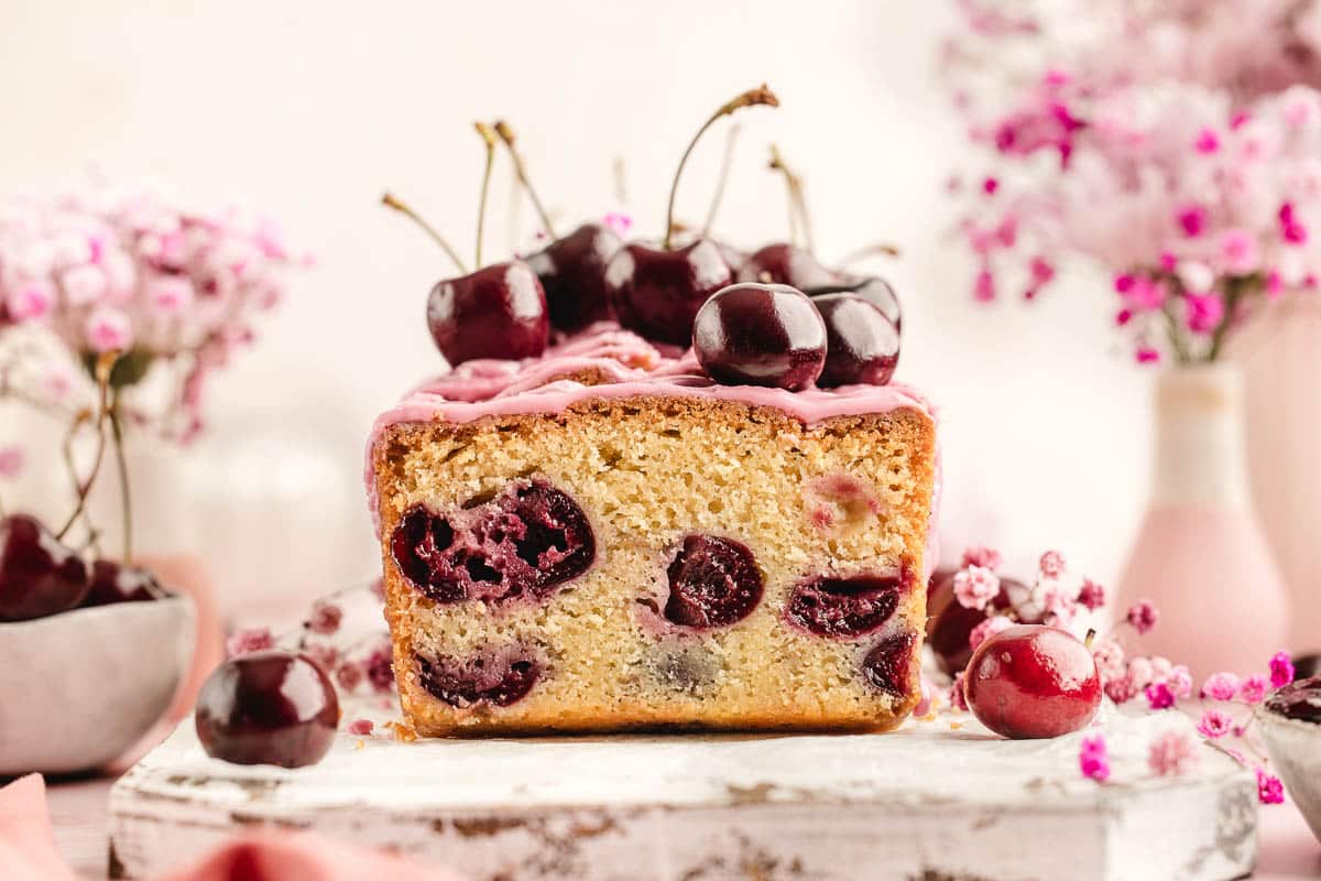 A loaf of cherry cake topped with fresh cherries and pink icing, with slices of cherries visible inside the cake and pink flowers in the background.