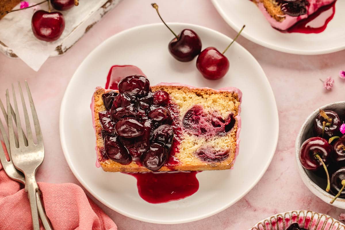A slice of cherry loaf cake topped with cherry compote on a white plate, with fresh cherries and a fork beside it.