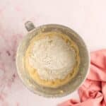A metal mixing bowl with flour and dough sits on a pink surface next to a folded pink cloth.