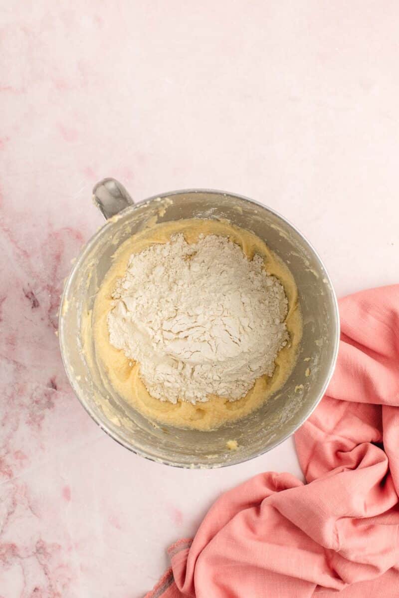 A metal mixing bowl with flour and dough sits on a pink surface next to a folded pink cloth.
