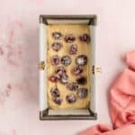 A loaf pan lined with parchment paper filled with batter and topped with sugared cranberries, placed on a pink marble surface next to a pink cloth.