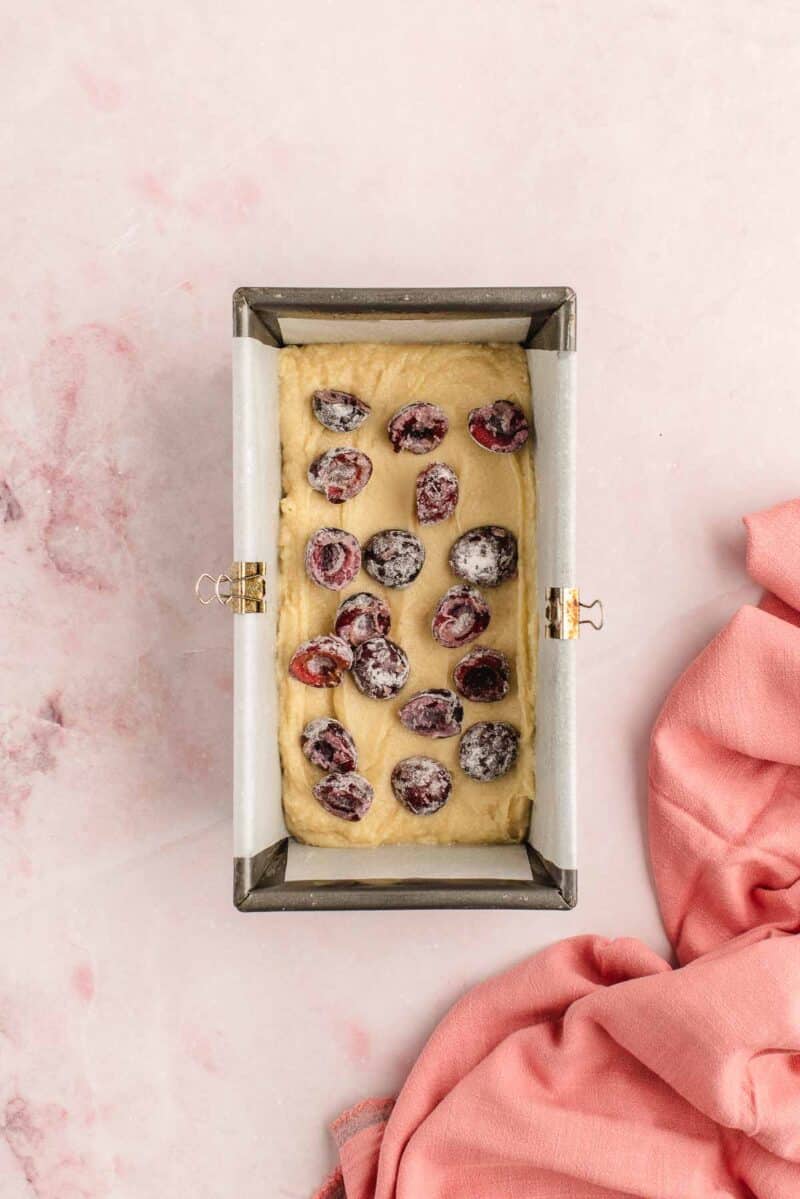 A loaf pan lined with parchment paper filled with batter and topped with sugared cranberries, placed on a pink marble surface next to a pink cloth.