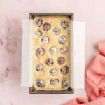 A rectangular loaf pan lined with parchment paper holds unbaked batter topped with scattered cherries, set on a light pink surface beside a pink cloth.