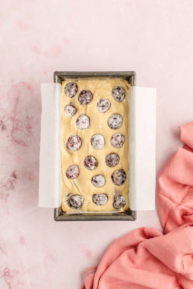 A rectangular loaf pan lined with parchment paper holds unbaked batter topped with scattered cherries, set on a light pink surface beside a pink cloth.