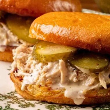 Close-up of two loaded sandwiches with shredded chicken, pickles, and white sauce on toasted buns, served on a decorative plate—perfect for better lunches or adding to your favorite lunch recipes.