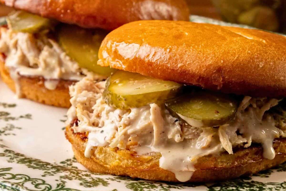 Close-up of two loaded sandwiches with shredded chicken, pickles, and white sauce on toasted buns, served on a decorative plate—perfect for better lunches or adding to your favorite lunch recipes.