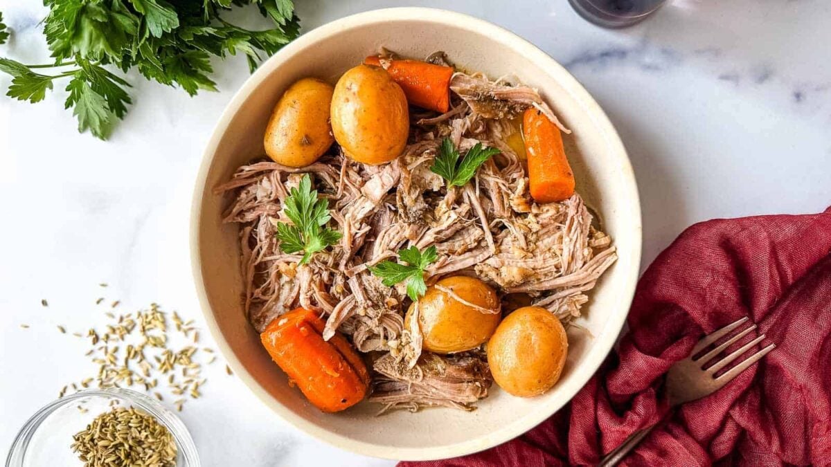 A bowl of shredded meat, whole baby potatoes, and carrot pieces garnished with parsley—an easy dinner for any night. A fork, parsley, and fennel seeds are nearby on the marble surface, perfect for fans of slow cooker pork recipes.