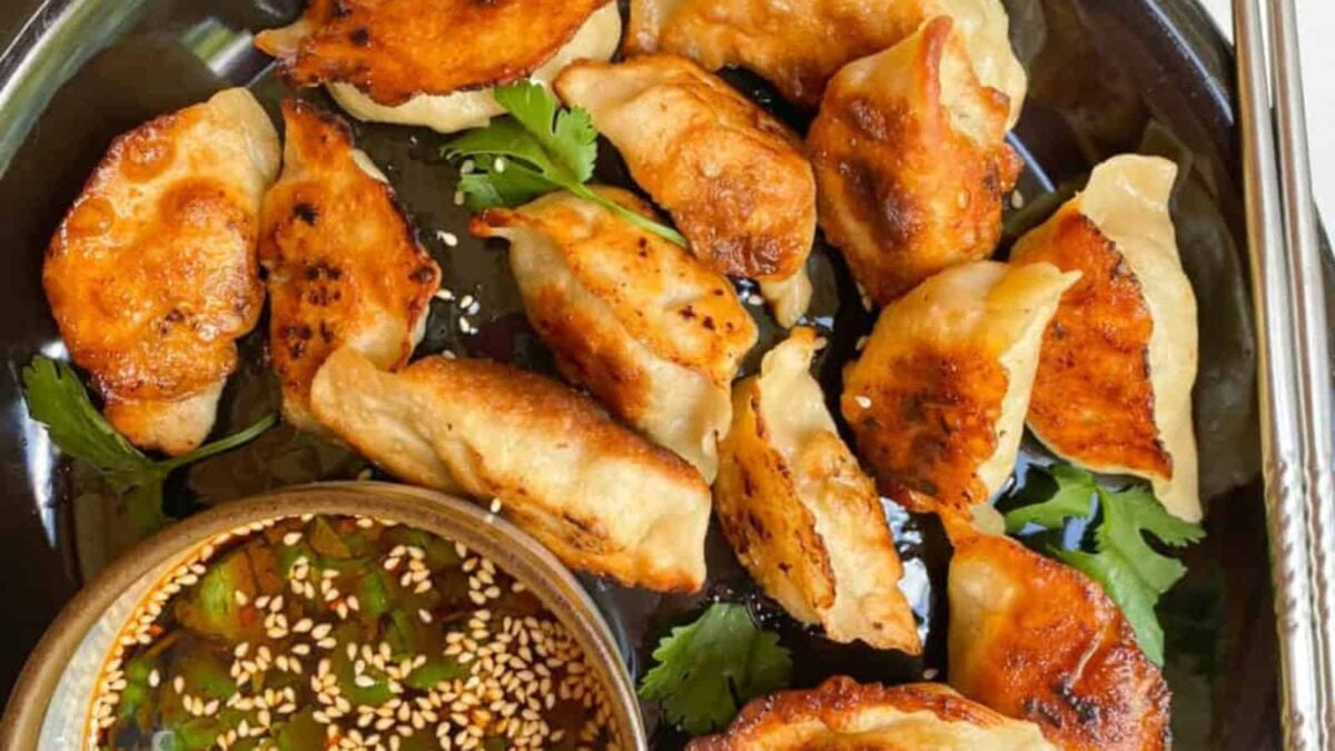 A plate of golden-brown pan-fried dumplings served with a bowl of soy dipping sauce topped with sesame seeds and fresh herbs—perfect for those craving handmade comfort and cozy recipes.