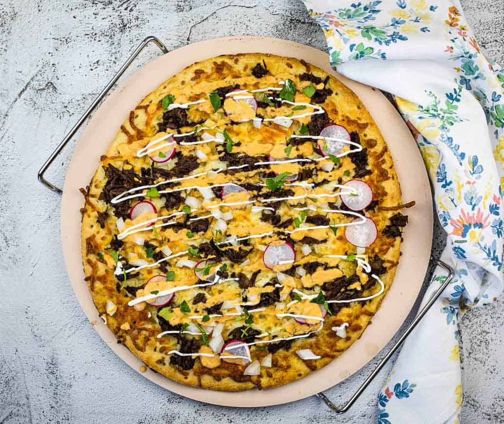 A fresh pizza topped with sliced radishes, herbs, shredded meat, cheese, and a drizzle of white sauce sits on a round tray beside a floral-patterned cloth—perfect for new pizza ideas on Friday nights.