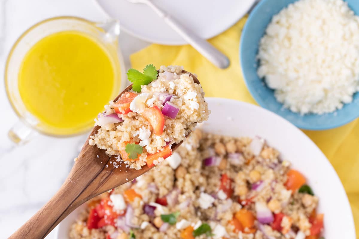 A wooden spoon holds a serving of quinoa salad with chopped vegetables and feta cheese above a bowl of the same salad, with a blue bowl of crumbled cheese and a glass container of yellow dressing nearby.