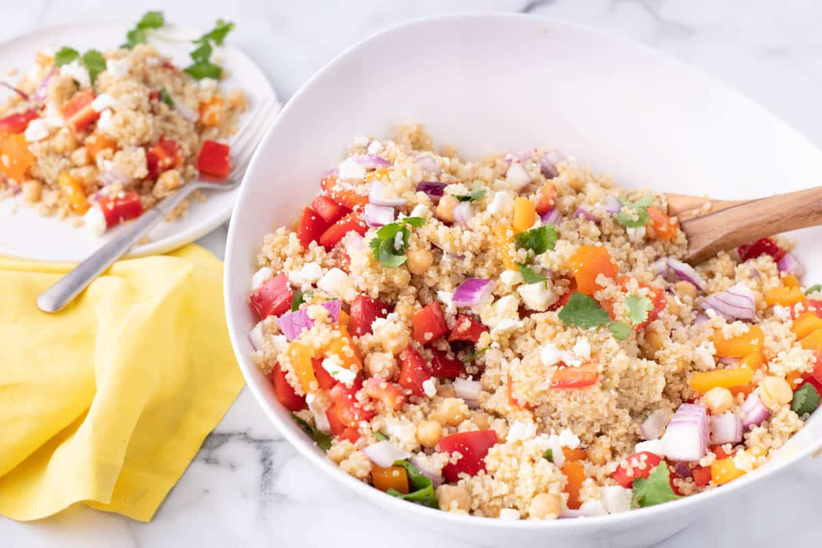 A bowl of quinoa salad with chopped red onions, bell peppers, chickpeas, feta cheese, and cilantro. A serving is on a plate with a fork and a yellow napkin beside it.