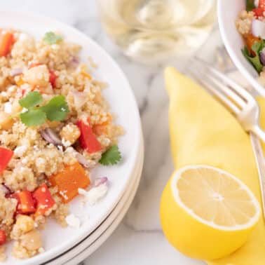 A plate of quinoa salad with red and orange bell peppers, chickpeas, red onion, cilantro, and feta cheese, next to a lemon half, cutlery, and a glass of white wine.