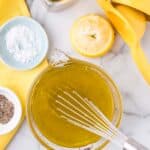 A glass bowl of yellow vinaigrette being whisked, with a halved lemon, yellow juicer, small bowls of salt and pepper, olive oil, and a yellow napkin on a marble surface.