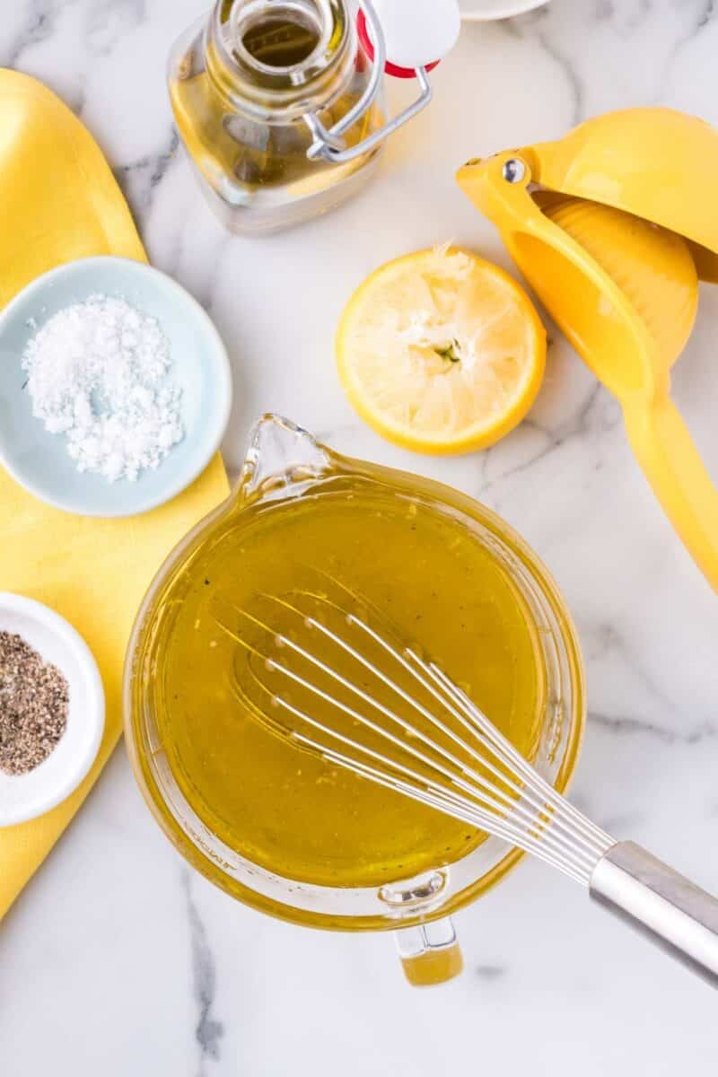 A glass bowl of yellow vinaigrette being whisked, with a halved lemon, yellow juicer, small bowls of salt and pepper, olive oil, and a yellow napkin on a marble surface.