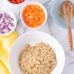 A bowl of cooked quinoa surrounded by small bowls of chopped red peppers, yellow peppers, red onions, and chickpeas on a marble surface with a wooden spoon.