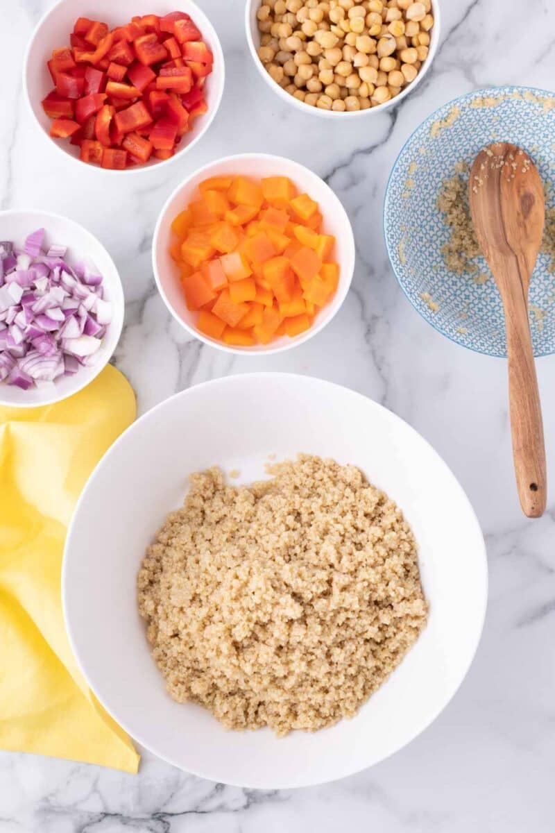 A bowl of cooked quinoa surrounded by small bowls of chopped red peppers, yellow peppers, red onions, and chickpeas on a marble surface with a wooden spoon.