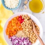 A bowl with chopped yellow and red bell peppers, red onion, quinoa, and chickpeas, next to a bowl of feta cheese and a cup of olive oil on a marble surface.