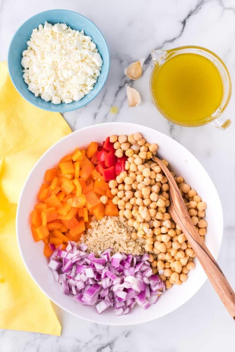 A bowl with chopped yellow and red bell peppers, red onion, quinoa, and chickpeas, next to a bowl of feta cheese and a cup of olive oil on a marble surface.