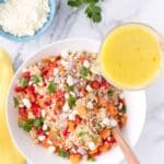 A bowl of quinoa salad with chopped vegetables and cheese is being topped with yellow dressing, with a side dish of crumbled cheese and cilantro on a marble surface.