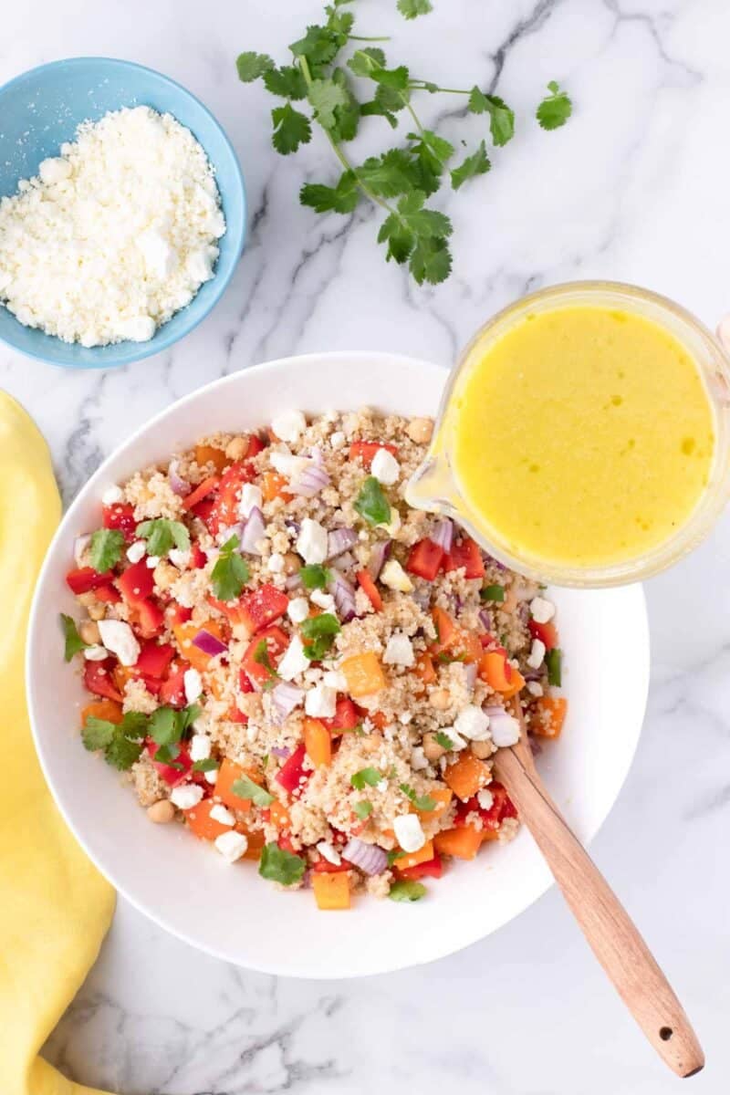 A bowl of quinoa salad with chopped vegetables and cheese is being topped with yellow dressing, with a side dish of crumbled cheese and cilantro on a marble surface.