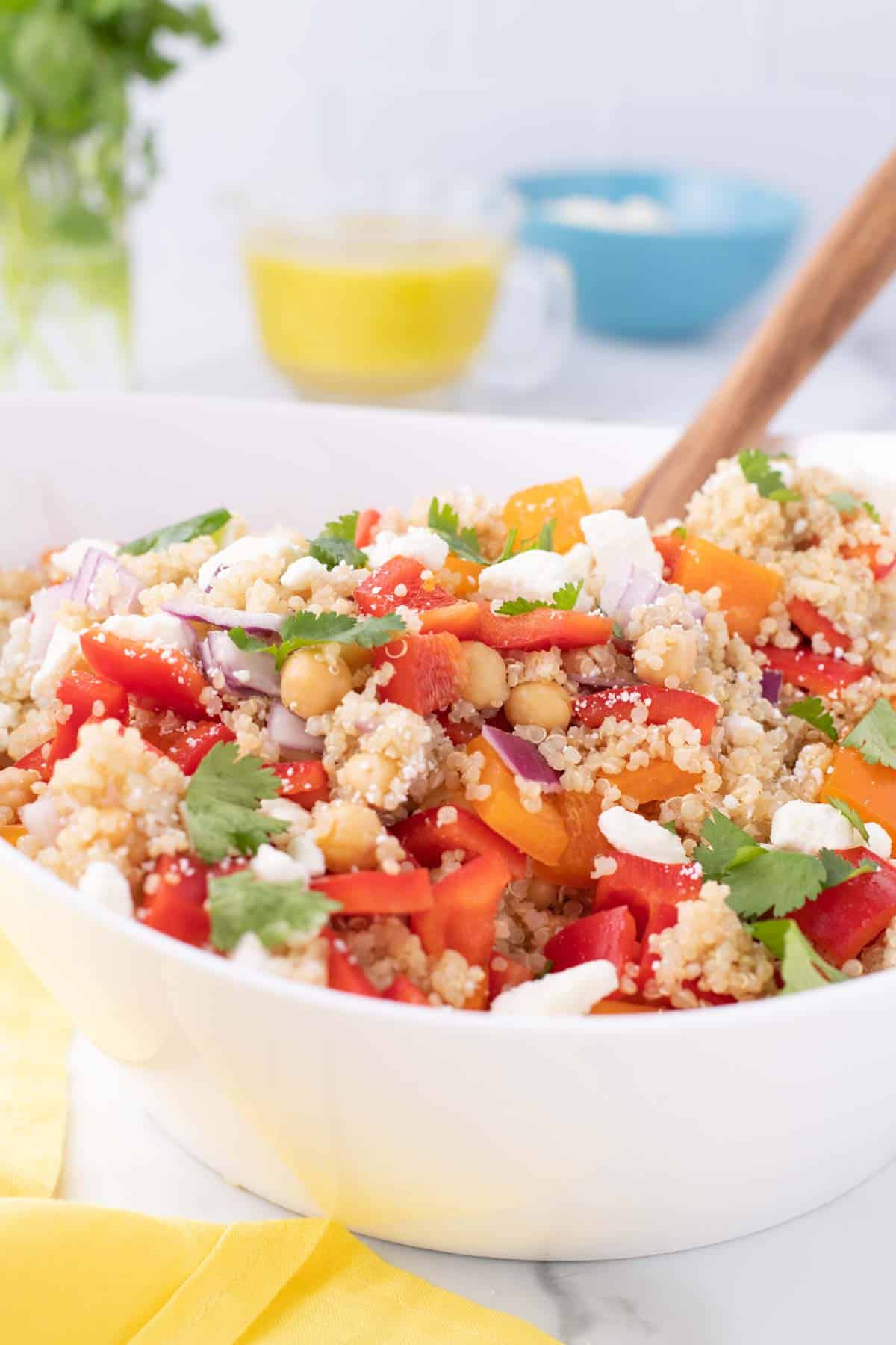 A white bowl filled with quinoa salad, featuring chopped red and yellow bell peppers, chickpeas, red onion, cilantro, and crumbled cheese, with a wooden spoon inside.