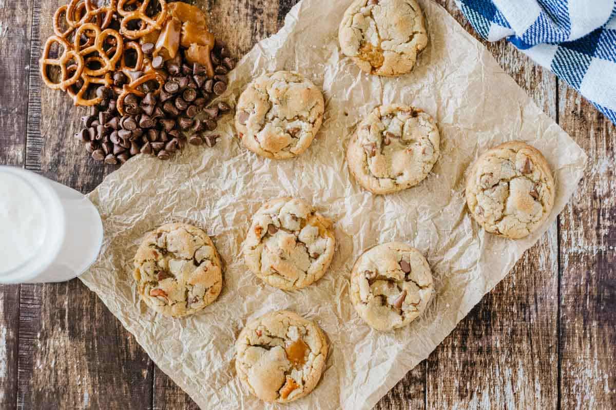 Eight cookies are arranged on crumpled parchment paper, with chocolate chips, pretzels, caramel pieces, and a glass of milk nearby on a wooden surface.
