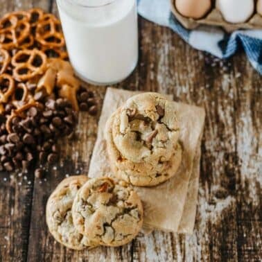 A stack of chocolate chip cookies on parchment paper next to pretzels, chocolate chips, a glass of milk, eggs, and a blue-checkered cloth on a rustic wooden surface.