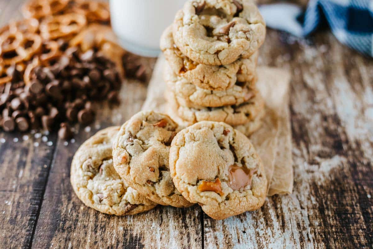 A stack of chocolate chip cookies with pretzels and chocolate chips scattered on a wooden surface, with a glass of milk in the background.
