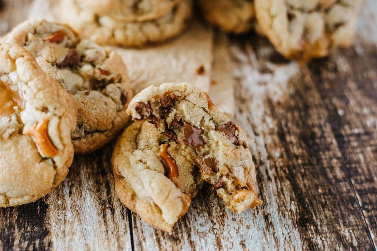 A close-up of chocolate chip cookies with caramel pieces, one cookie broken in half, on a rustic wooden surface.