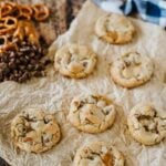 Seven cookies with visible pretzel and chocolate pieces are arranged on crumpled parchment paper, with pretzels, chocolate chips, caramel, and eggs in the background.