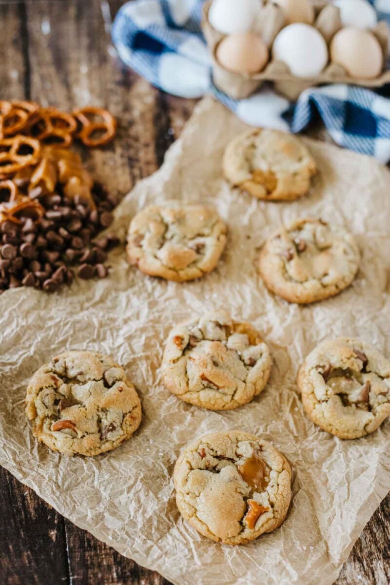 Seven cookies with visible pretzel and chocolate pieces are arranged on crumpled parchment paper, with pretzels, chocolate chips, caramel, and eggs in the background.
