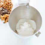 A stand mixer bowl with sugar and shortening inside, next to pretzels, chocolate chips, and caramel pieces on a white surface.