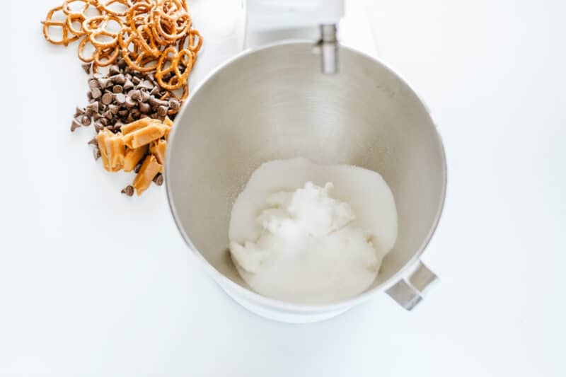 A stand mixer bowl with sugar and shortening inside, next to pretzels, chocolate chips, and caramel pieces on a white surface.