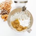 A stand mixer bowl with sugar, brown sugar, and butter being mixed, next to pretzels, caramel pieces, and chocolate chips on a white surface.