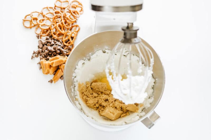A stand mixer bowl with sugar, brown sugar, and butter being mixed, next to pretzels, caramel pieces, and chocolate chips on a white surface.
