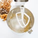 A stand mixer with partially mixed dough and flour in the bowl. Nearby are pretzels, chocolate chips, caramel pieces, and a sifter with flour.