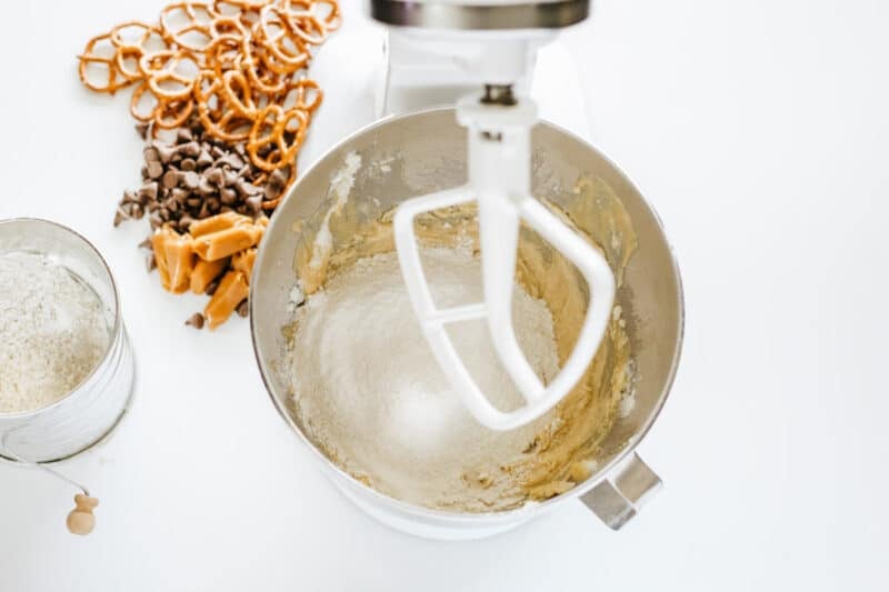 A stand mixer with partially mixed dough and flour in the bowl. Nearby are pretzels, chocolate chips, caramel pieces, and a sifter with flour.