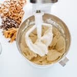 A stand mixer with cookie dough, surrounded by flour, chocolate chips, caramel pieces, and pretzels on a white surface.