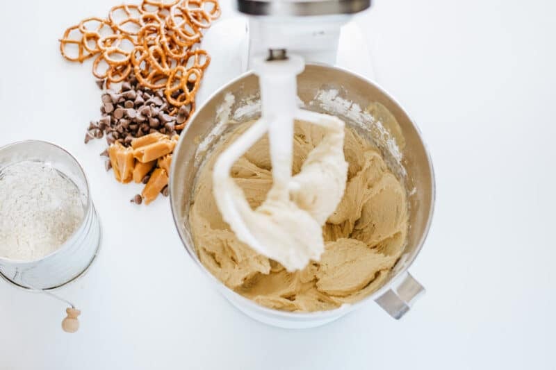 A stand mixer with cookie dough, surrounded by flour, chocolate chips, caramel pieces, and pretzels on a white surface.