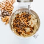 A stand mixer bowl containing cookie dough mixed with pretzel pieces and chocolate chips, with extra pretzels, caramel pieces, and chocolate chips beside the bowl.