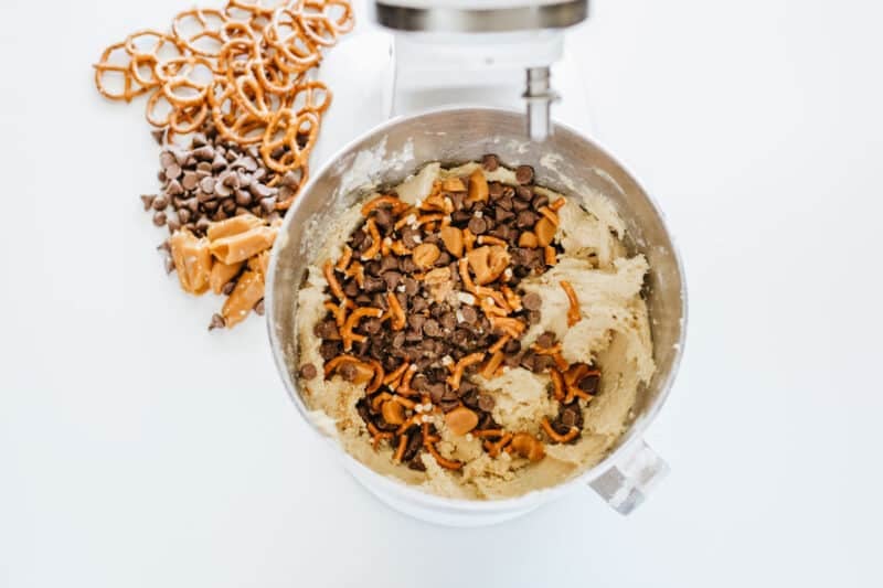A stand mixer bowl containing cookie dough mixed with pretzel pieces and chocolate chips, with extra pretzels, caramel pieces, and chocolate chips beside the bowl.