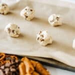 Balls of cookie dough with chocolate chips spaced on a parchment-lined baking sheet, with pretzels and chocolate chips visible in the foreground.