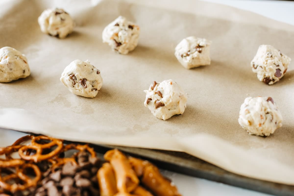 Balls of cookie dough with chocolate chips spaced on a parchment-lined baking sheet, with pretzels and chocolate chips visible in the foreground.