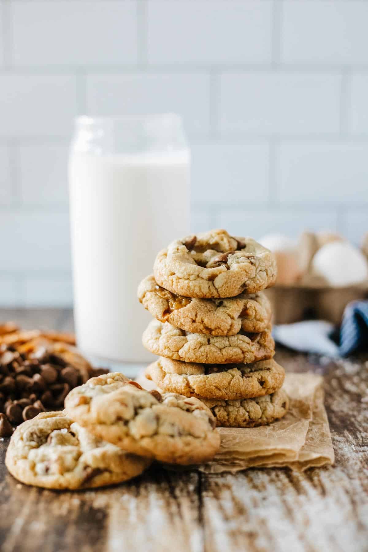 A stack of chocolate chip cookies sits on parchment paper in front of a glass of milk, with eggs and chocolate chips in the background.