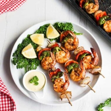Grilled shrimp and vegetable skewers served on a white plate with lemon wedges, parsley, and dipping sauce; perfect for weeknight meals or when you need quick seafood recipes. Grilling tray and red checkered cloth nearby.