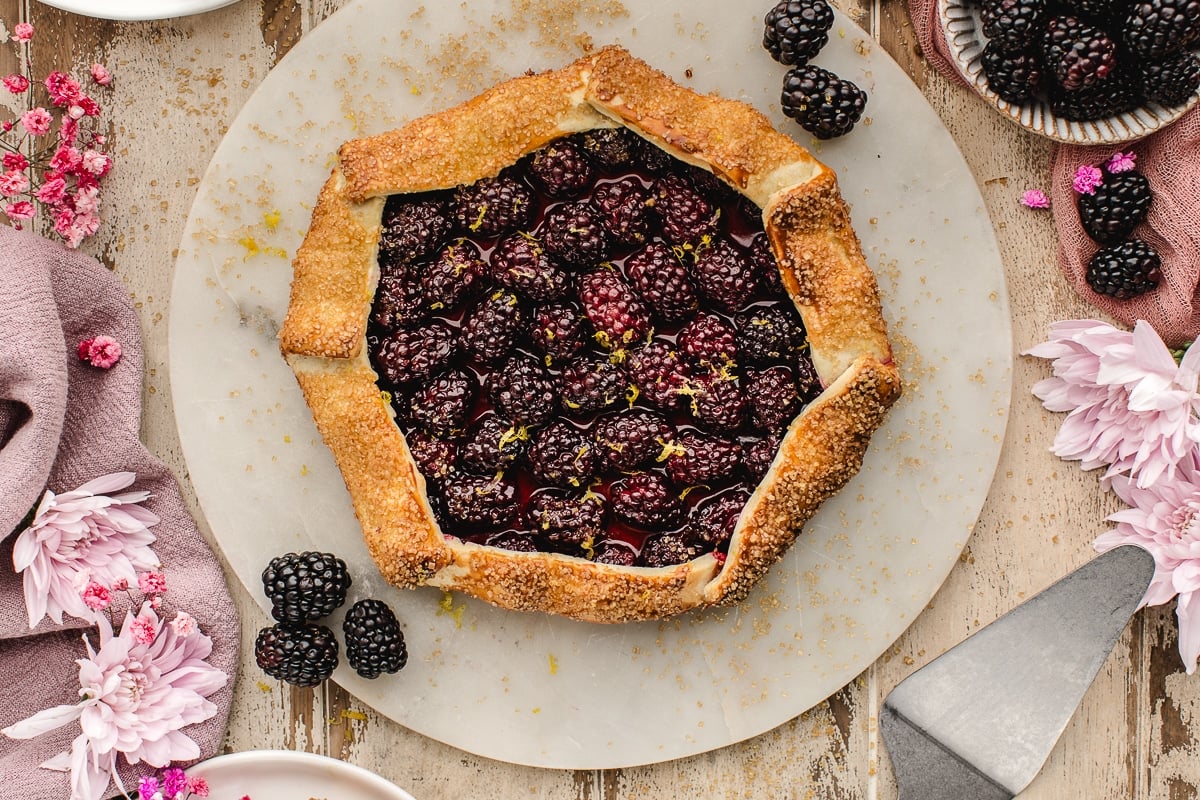 A rustic blackberry galette with a golden crust sits on a round marble board, surrounded by fresh blackberries and pink flowers.