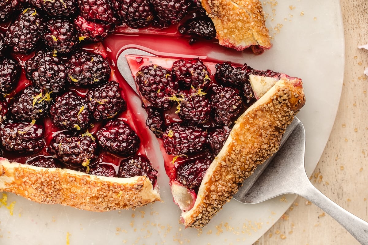 A close-up of a blackberry galette with a slice being lifted on a metal spatula, showing a golden crust and juicy berries.
